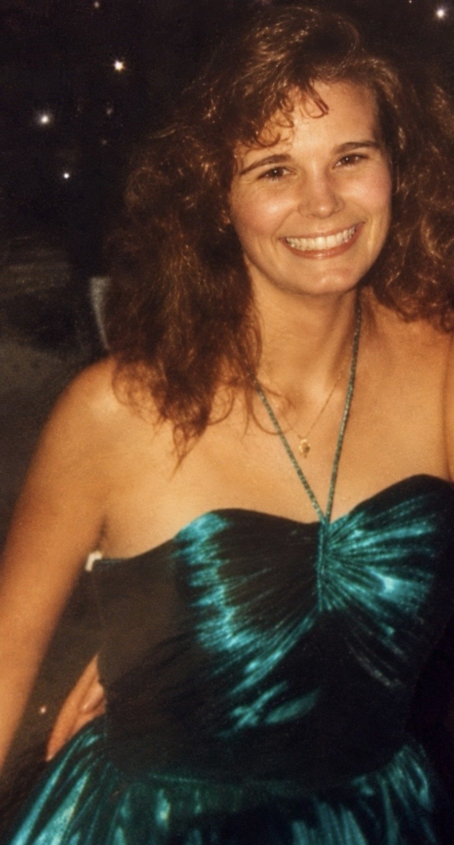 A woman with curly reddish-brown hair, smiling, wearing a strapless black dress with teal accents, and a necklace with a small pendant.