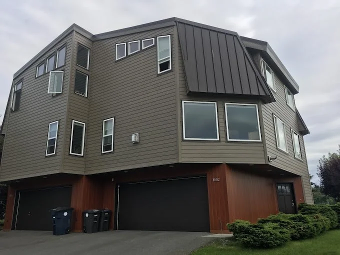Modern three-story house with gray siding, numerous windows, and a dark metal roof, with garage doors on the ground level and trash bins outside.