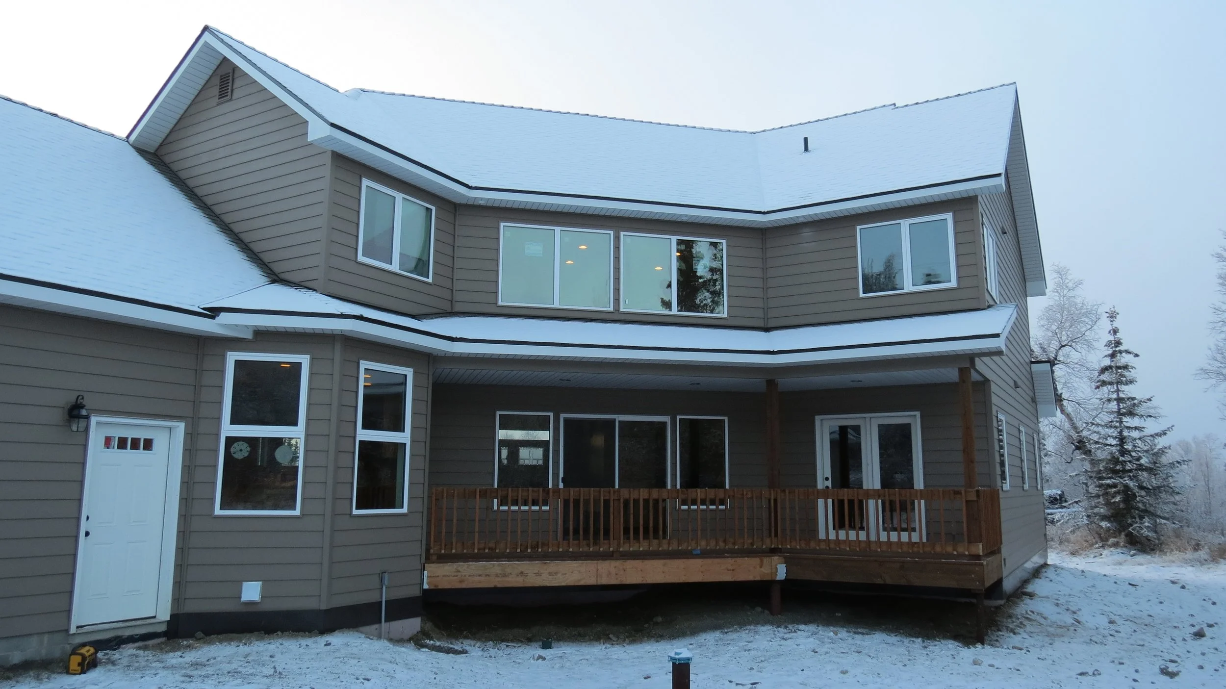 A two-story house with beige siding, multiple windows, and a snow-covered roof, with a deck and a wooden fence in the backyard, set in a cold or wintery environment.