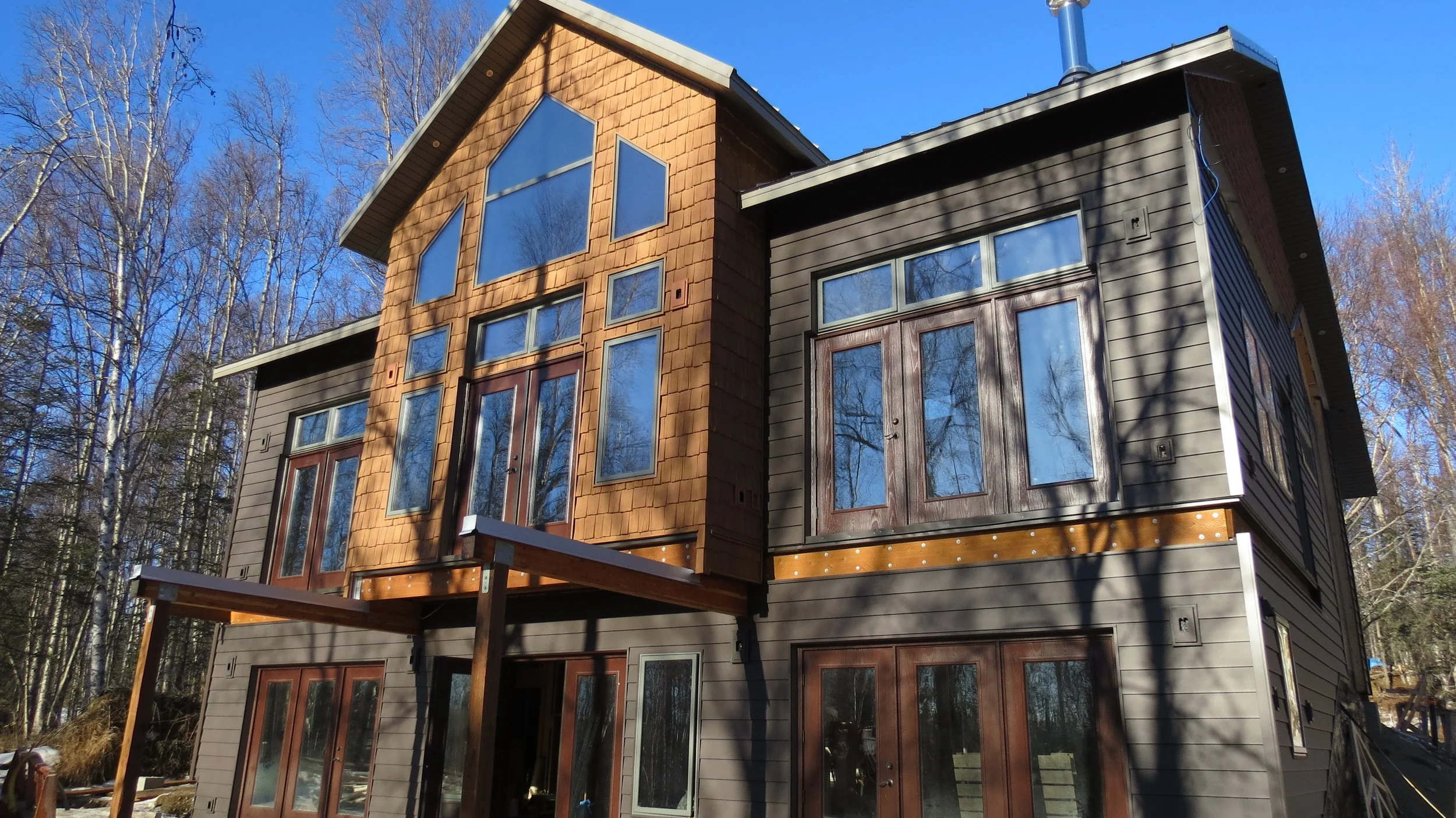 Modern multi-story house with large windows, brown and gray siding, situated in a wooded area with bare trees, under a clear blue sky.