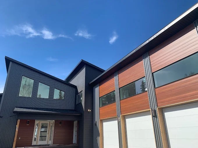 Modern house with black and wooden exterior, large windows, and a blue sky with a few clouds.