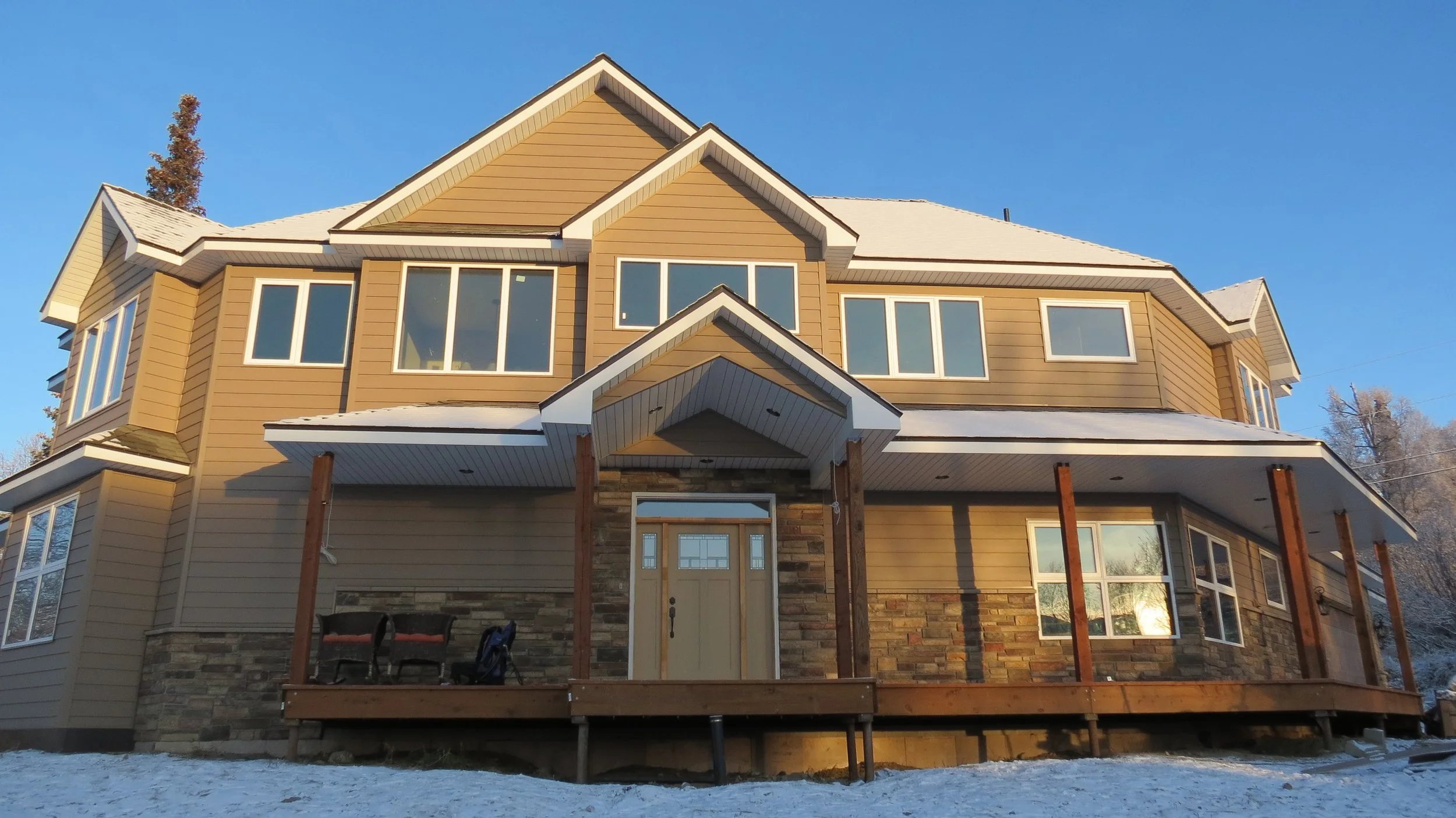 A large multi-story house with beige and brown siding, multiple windows, a stone facade at the lower level, and an unfinished wooden deck in the front yard. Snow is on the ground and the sky is clear and blue.