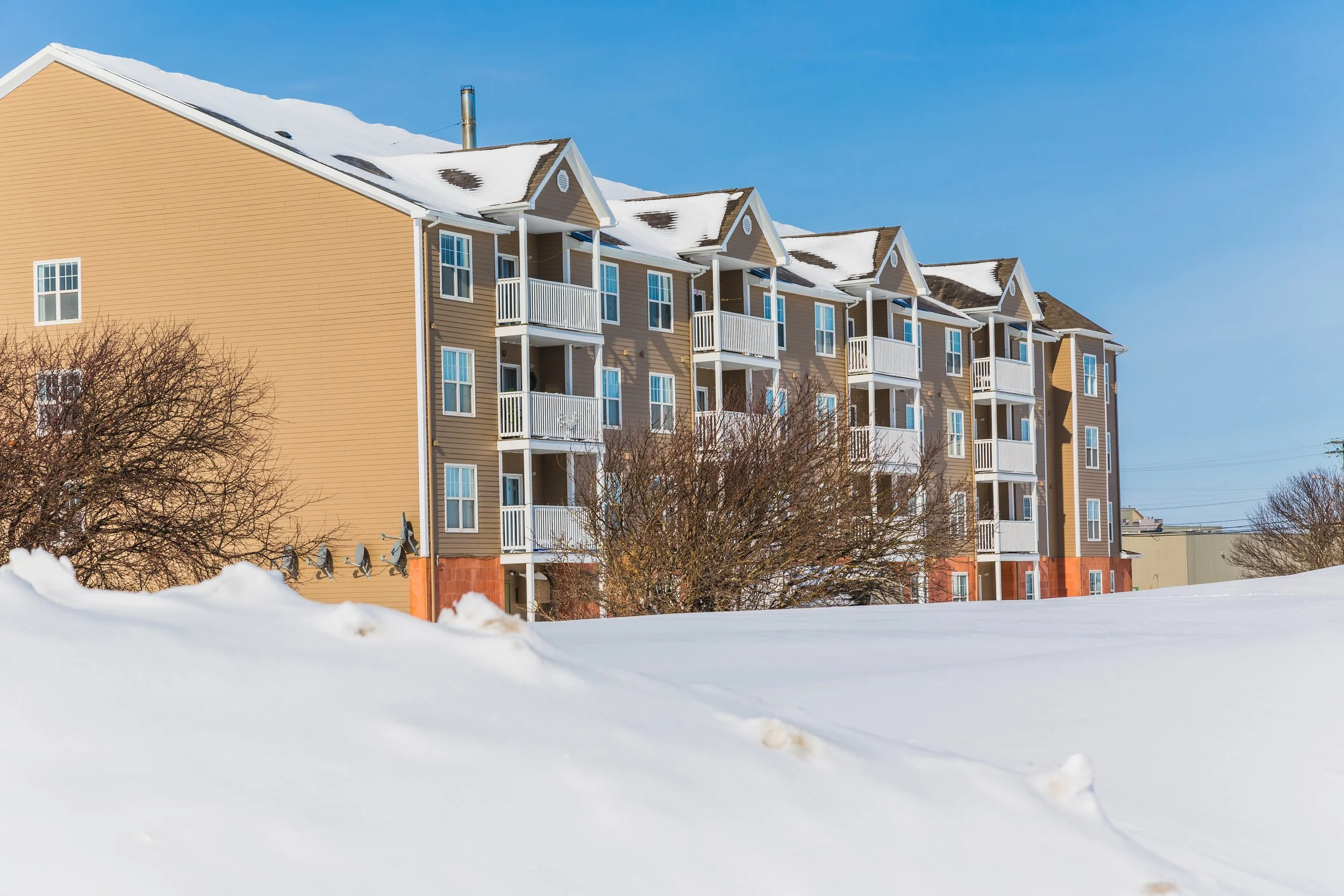 Multistory apartment building with beige siding and white balconies in winter, snow on roof and ground, against a blue sky.