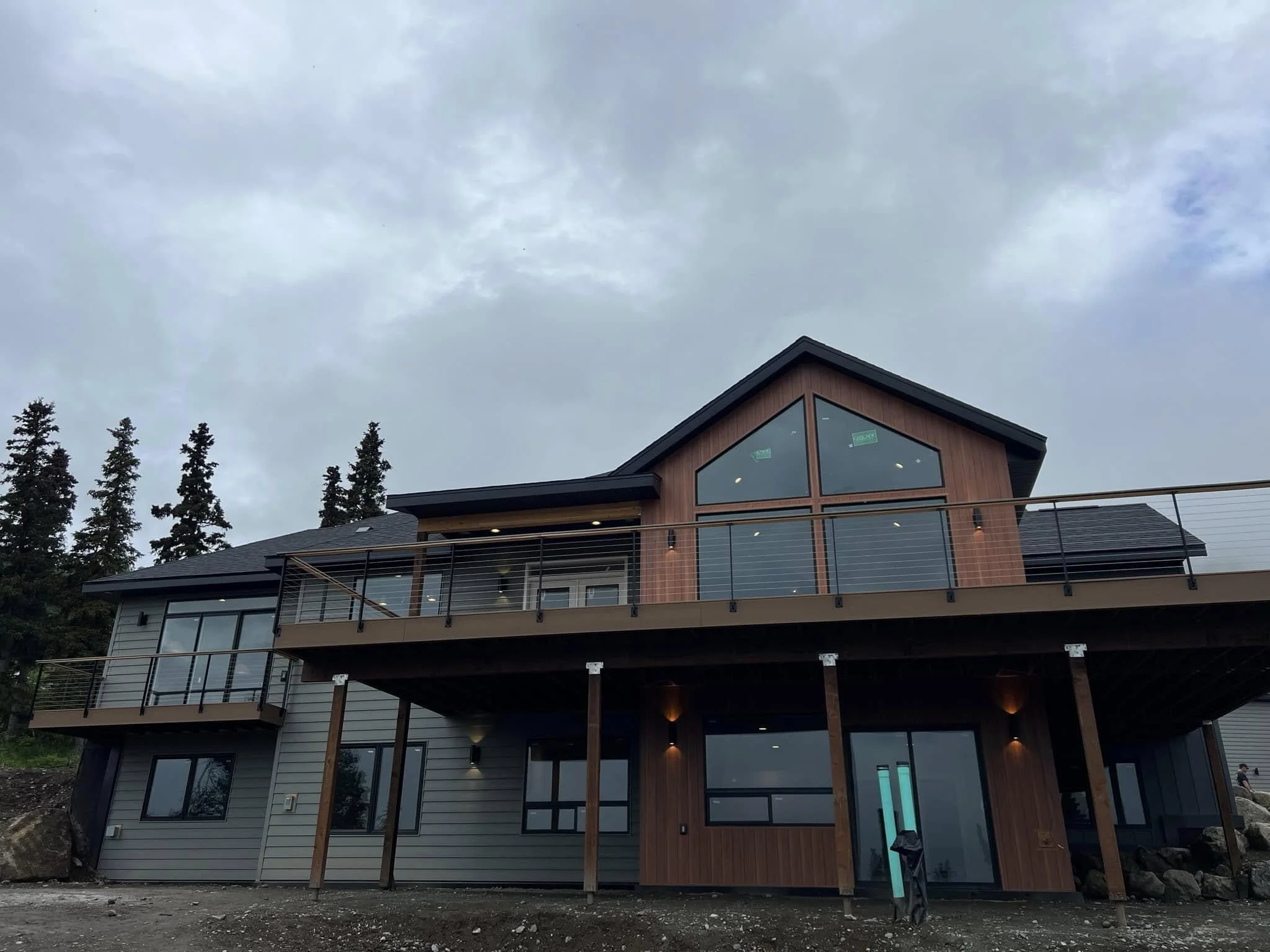 Modern two-story house with large windows, wooden and gray exterior, balcony, and surrounded by tall trees, under an overcast sky.