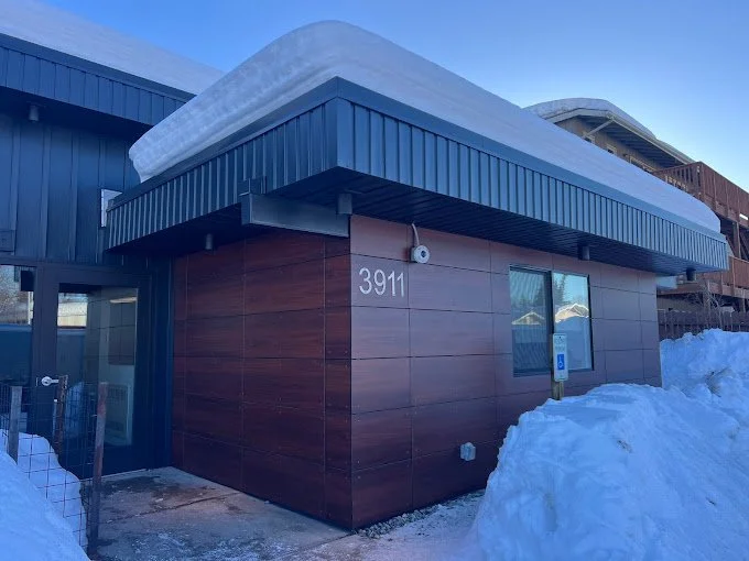 A modern building with a brown tiled facade, blue awning, and snow on the roof and ground, with the number 3911 displayed.