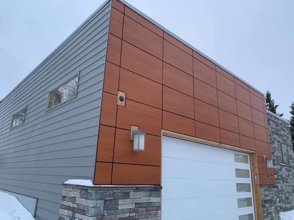 Modern house with gray and wood siding, stone accents, and a white garage door, amid snow and trees in the background.