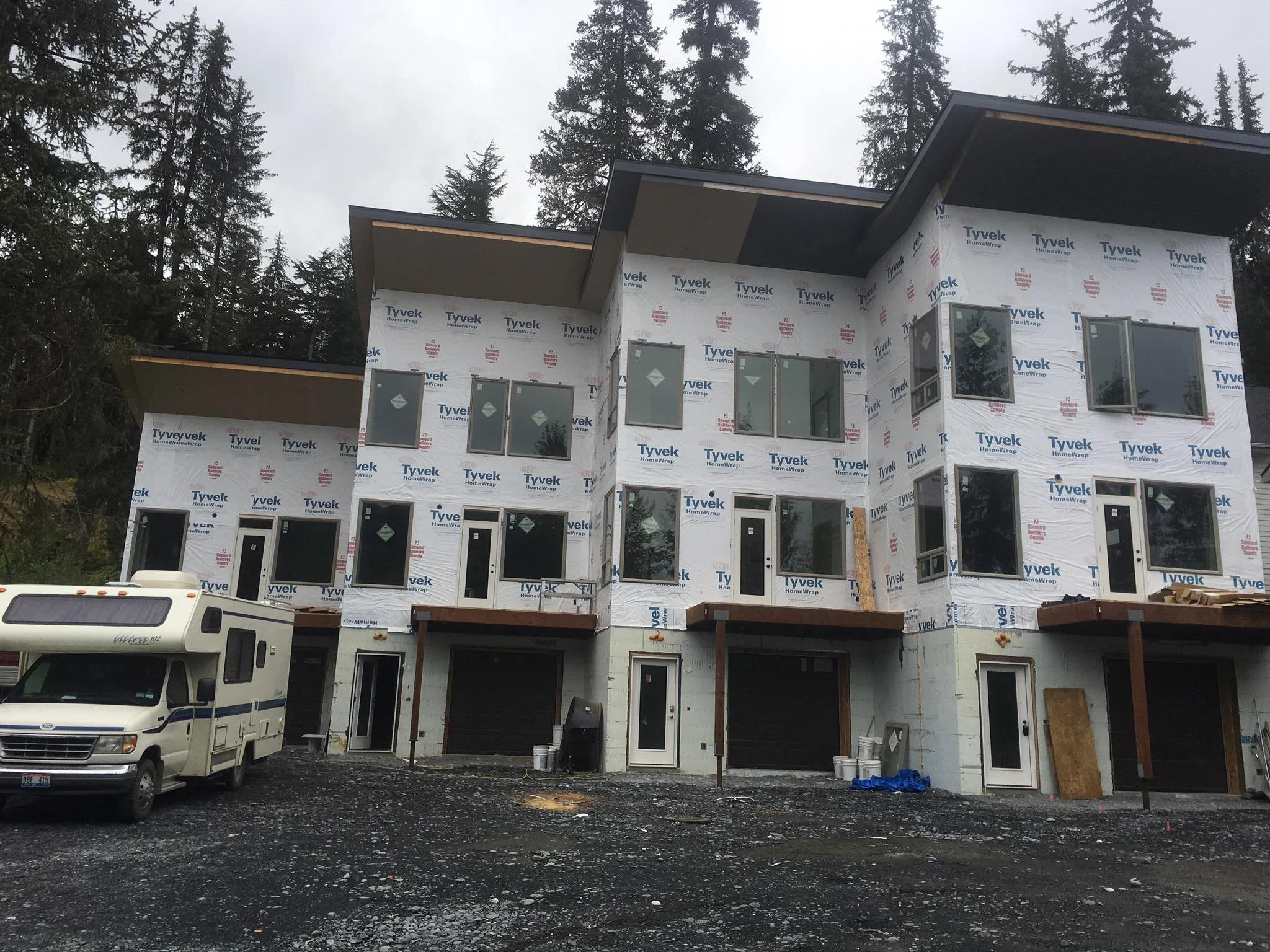 A multi-story house under construction, wrapped in Tyvek HomeWrap, surrounded by tall pine trees, with a white van and construction trailer in the foreground.