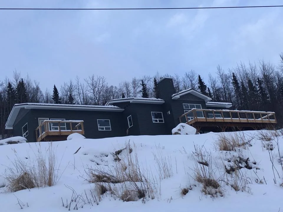 A modern black house on a snowy hillside with two wooden decks and leafless trees in the background.
