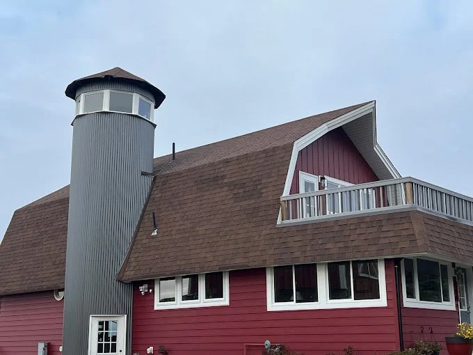 A red house with a brown shingle roof and a tall, round, gray metal chimney. The house has multiple windows and a small balcony on the upper level. The sky is overcast.