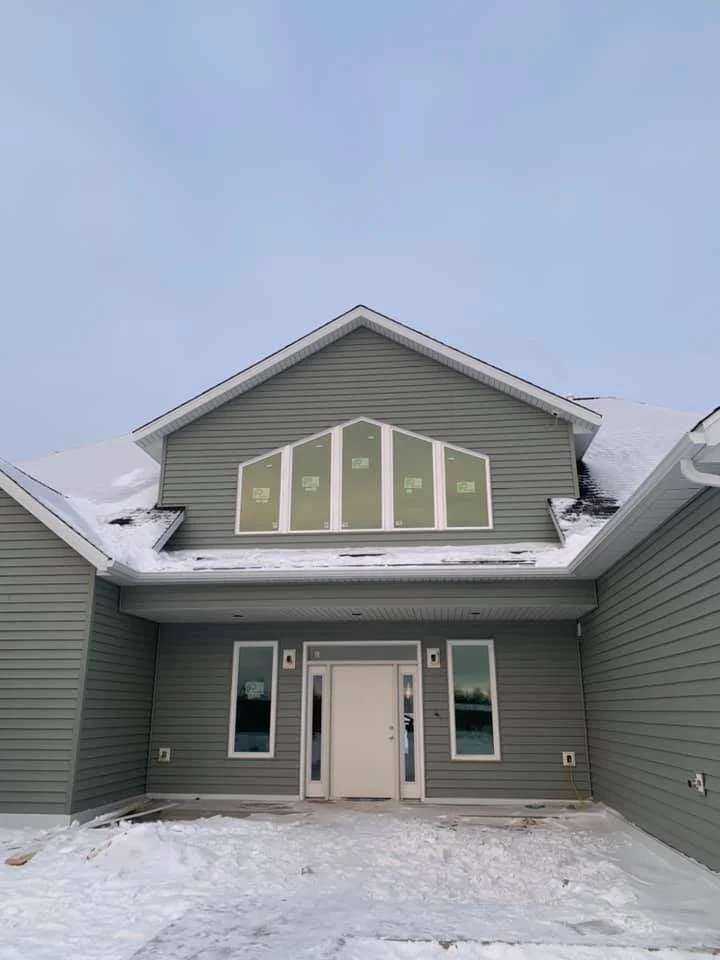 Front view of a new gray house with a snow-covered ground and roof, featuring a gabled roof, three windows on the upper level, and a single front door with side windows.