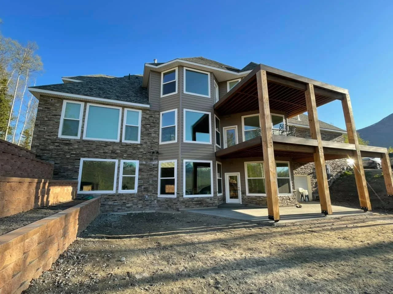 Newly constructed house with stone and siding exterior, multiple large windows, and a wooden deck with two levels in a mountainous area under a clear blue sky.