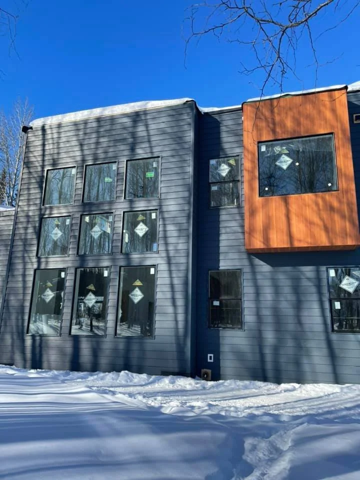 A modern multi-story building with gray and orange exterior siding, large windows, and snow on the ground and roof in winter.