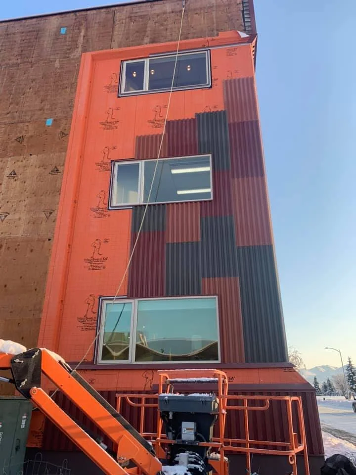 A multi-story building under construction with exterior panels that resemble a pixelated pattern in shades of orange, black, and dark red. There are three visible windows containing interior lighting, and a construction lift is at the bottom of the building.