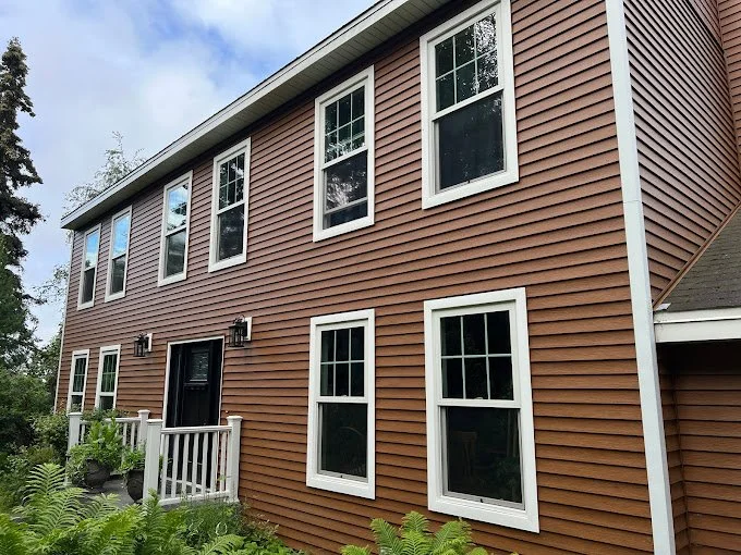 Front exterior view of a two-story house with brown horizontal siding, six double-hung windows, a black front door, and a small porch with white railings, surrounded by greenery.
