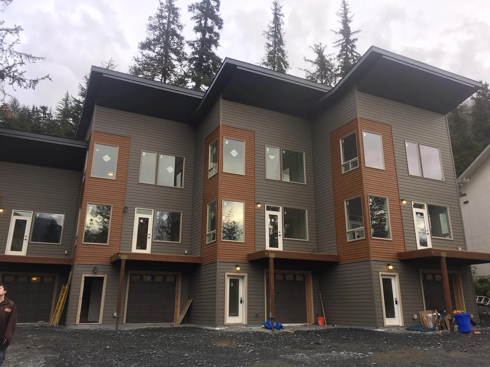 A modern multi-story residential building under construction, with gray and brown siding, numerous windows, and scaffolding or supports at the ground level, set against a backdrop of tall trees and a cloudy sky.