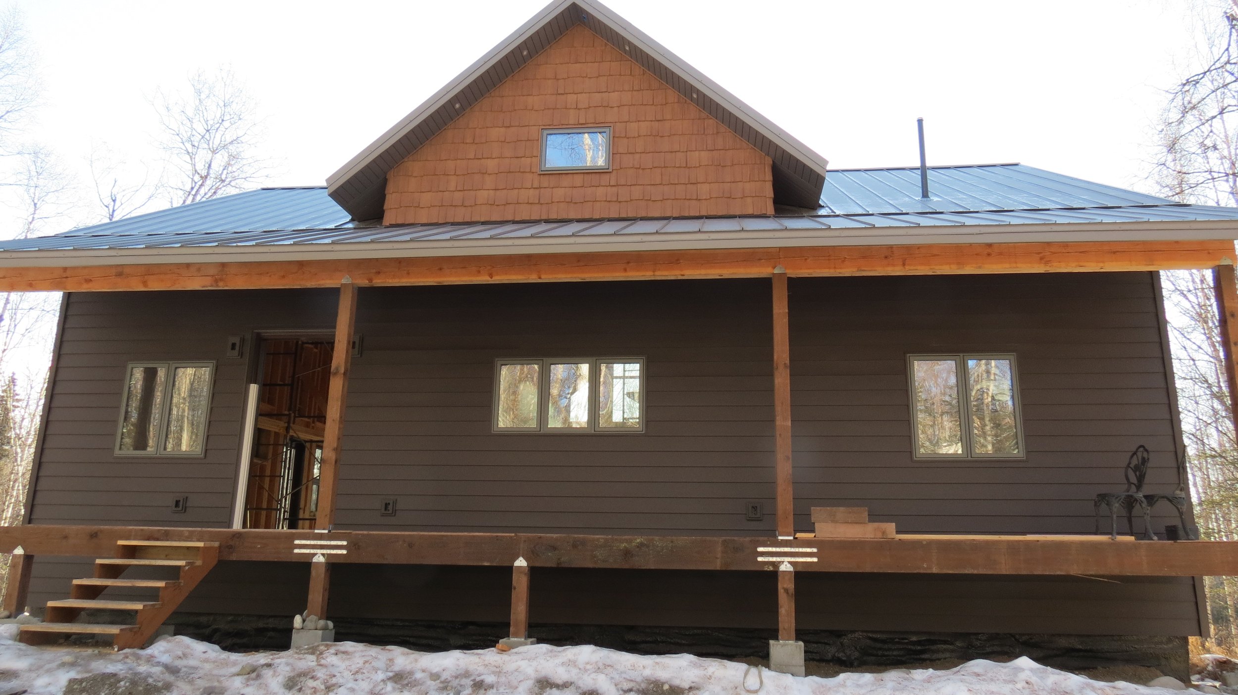 Back view of a two-story house under construction with a wooden deck and staircase, gray siding, and a gabled roof in a wooded area with snow on the ground.