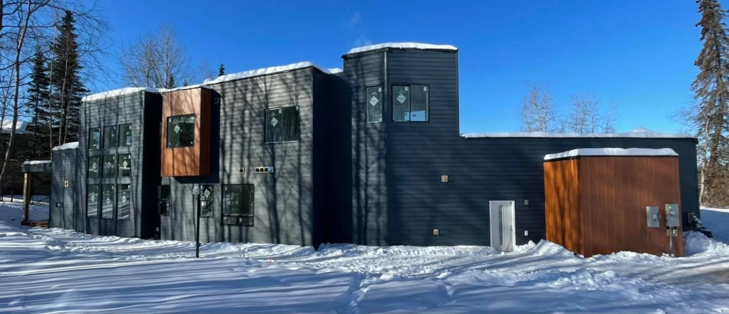 Modern multi-story house with dark siding, wooden accents, and snow-covered roof, surrounded by leafless trees against a bright blue sky.