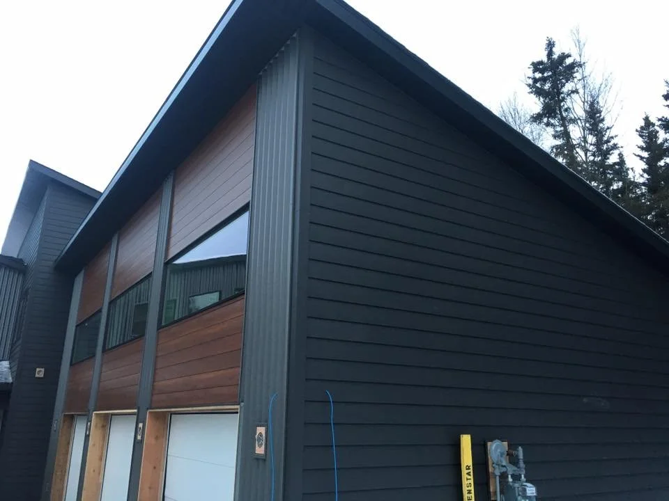 Front corner of a modern, multi-story building with black and wood panel siding, large diagonal windows, and a garage door. Tall trees are in the background.