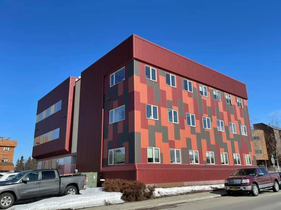 Multi-story building with a red and patterned exterior, parked cars below, and a clear blue sky.