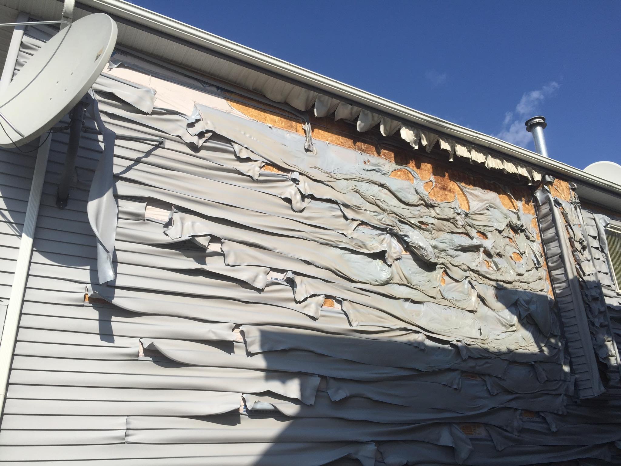 The image shows the side of a house with damaged and peeling siding, exposing the underlying insulation and wall structure. There are satellite dishes attached to the house and a metal chimney pipe is visible. The sky is clear and blue.