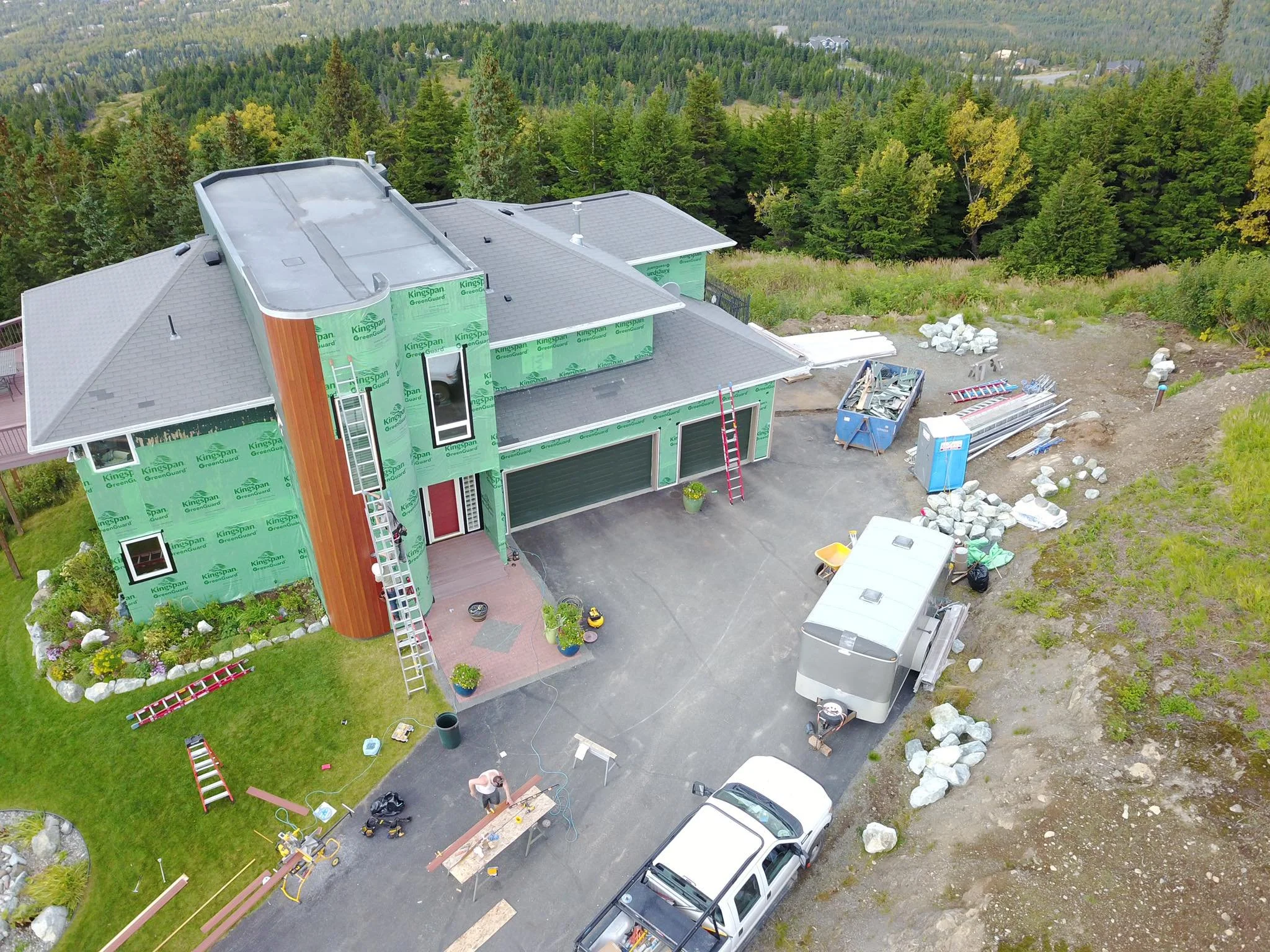 An aerial view of a house under construction surrounded by trees, with construction materials, tools, and vehicles on the driveway and yard.