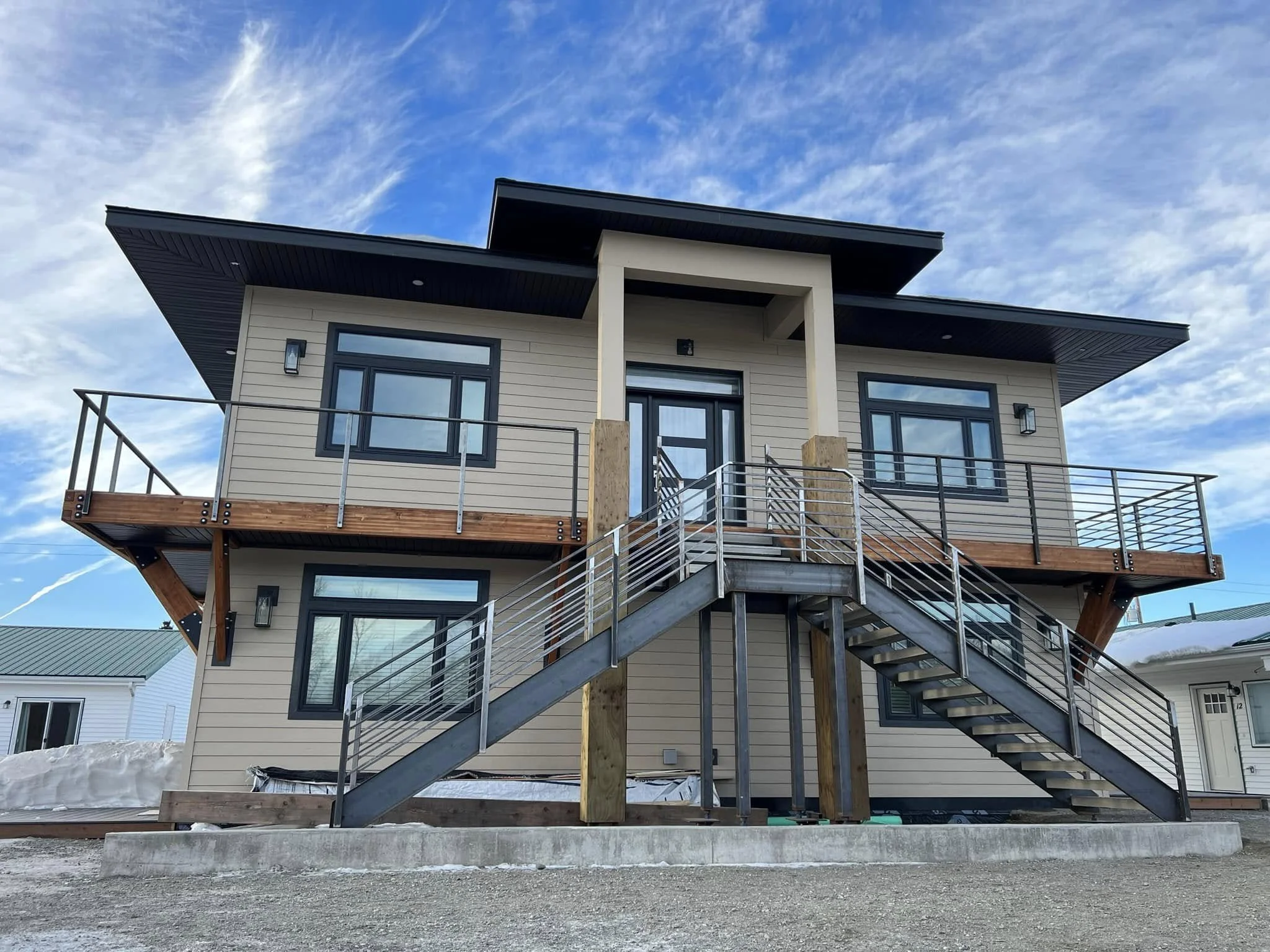 A modern, multi-story house under construction, with exterior stairs, large windows, and a roof with sharp angular slopes, against a blue sky with scattered clouds.