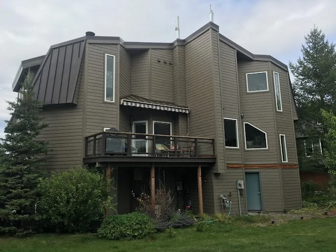 A multi-story house with angular design, gray siding, and large windows, surrounded by green trees and lawn.