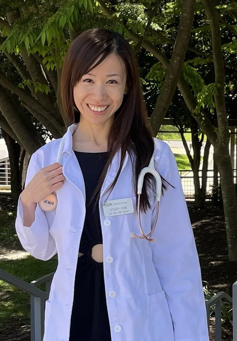 A smiling woman with long dark hair, wearing a white medical coat with a stethoscope around her neck and a name tag, standing outdoors near trees.