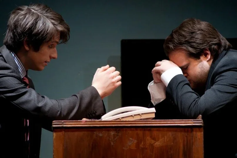 A man in a suit with long hair is sitting at a wooden desk, covering his face, appearing distressed. Another man in a suit is ssitting across from him, gesturing with his hand, in what seems to be a serious conversation.