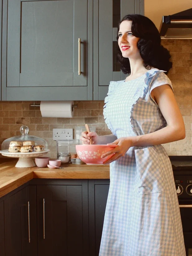 A woman with dark hair and red lipstick wearing a blue and white gingham apron standing in a modern kitchen, holding a mixing bowl and spoon, with cookies on a cake stand and baking ingredients on the countertop.