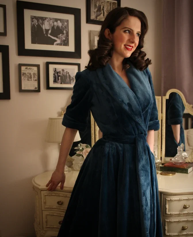 Woman with dark brown hair in vintage style, wearing a dark blue velvet dress, standing in a decorated bedroom with framed photos on the wall and a cream-colored dresser with mirror.