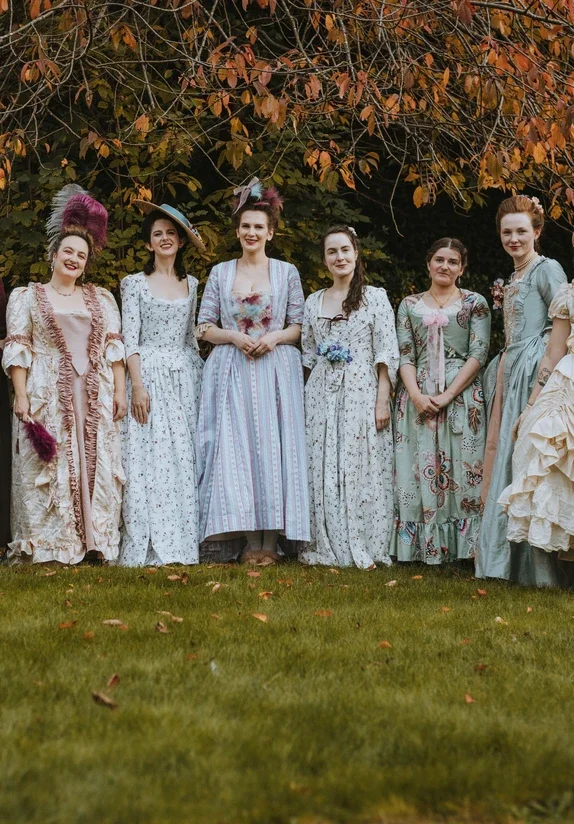 Six women dressed in Georgian dresses standing outdoors in front of trees with autumn leaves.