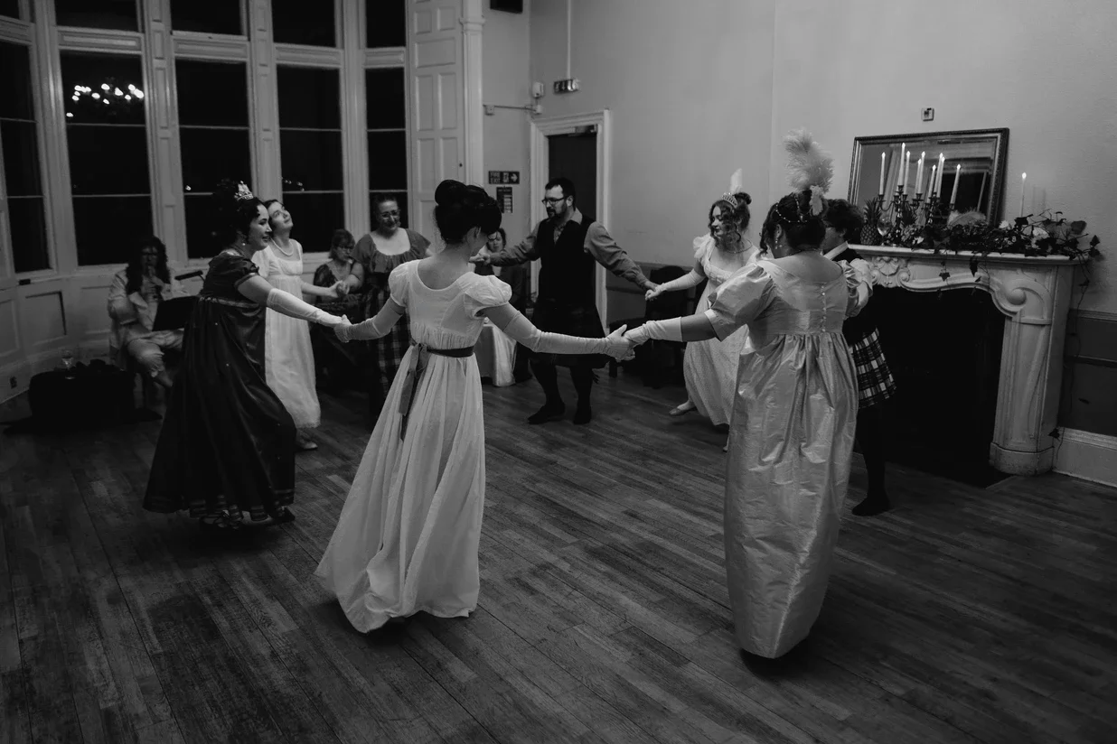 People in Regency costumes dancing in a circle during a celebration in a decorated room.