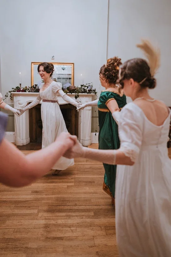 Women holding hands and dancing in a circle at a celebration, with a fireplace and mirror in the background.