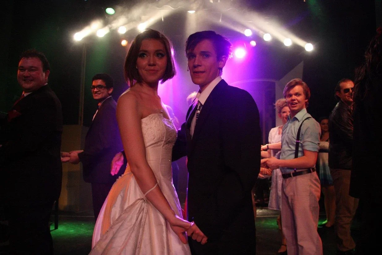 A couple dressed in formal attire dancing at a dance event, with other couples dancing and colorful stage lighting in the background.