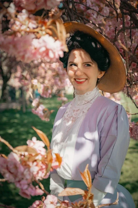 A woman wearing a wide-brimmed straw hat and Edwardian dress standing among pink cherry blossom trees.