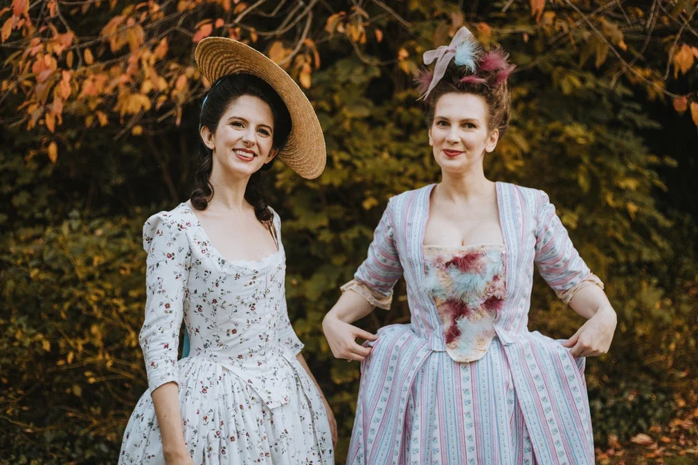 Two women dressed in floral Georgian gowns outdoors with autumn foliage in the background.