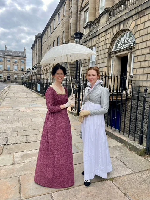 Two women dressed in historical clothing standing on a street in New Town, Edinburgh. One is holding a cream parasol, in front of a stone building with black iron fencing.