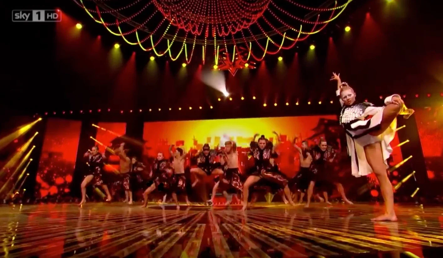 Dance performance on stage with multiple dancers, colorful lighting, and a bright background with cityscape imagery.
