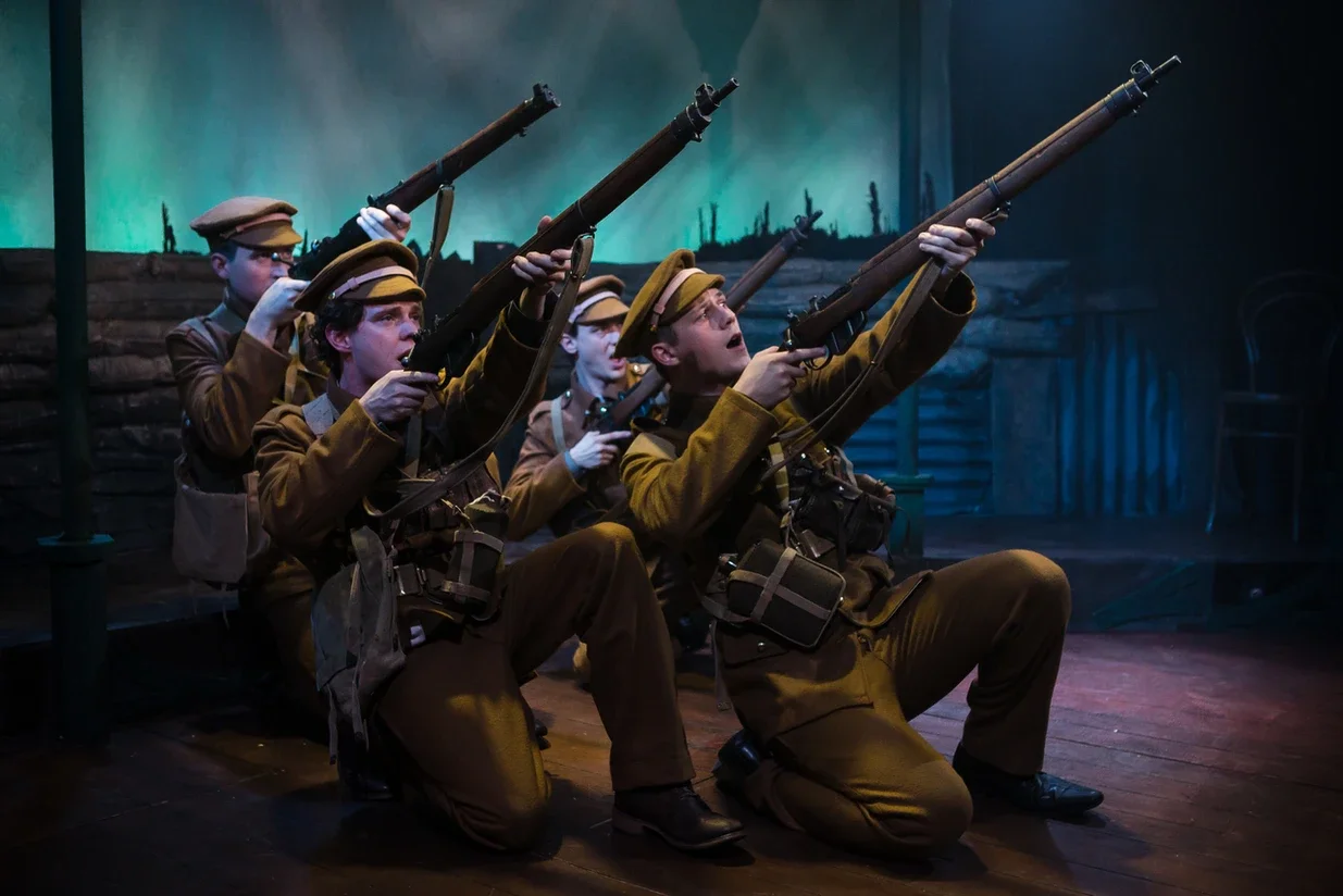 Group of soldiers in uniform kneeling and aiming rifles during a performance, with a dark background and dramatic lighting.