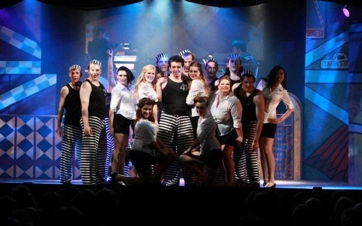 Group of performers on stage, dressed in black and white striped costumes, posing for a photo in front of a colorful stage backdrop.