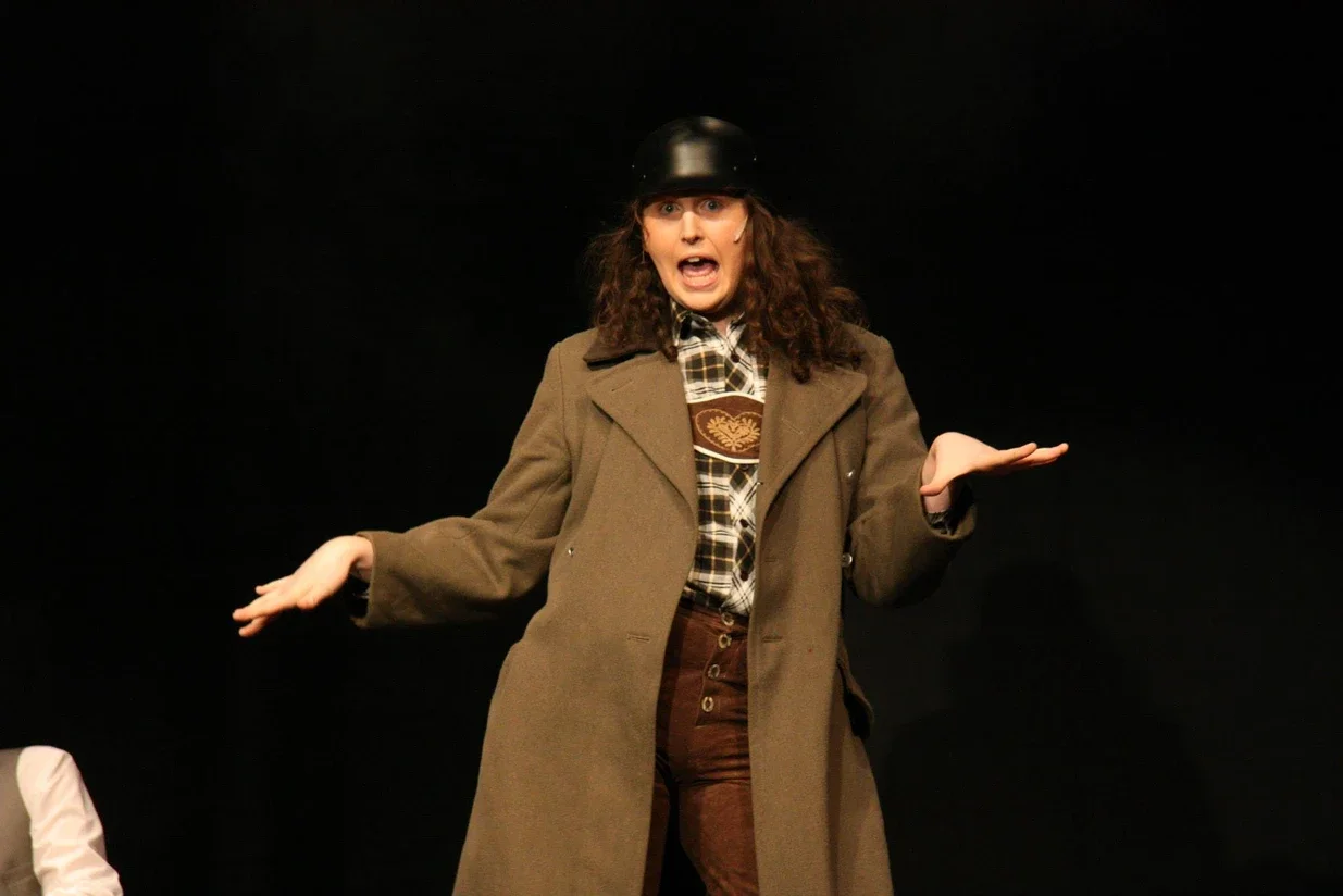 A man performing on stage, wearing a brown coat, plaid shirt, a black helmet, and brown pants, with his arms outstretched and a surprised or expressive facial expression against a black background.