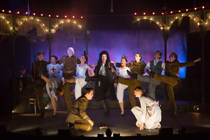 The image shows a theatrical stage scene with multiple actors in period costumes. Some are standing, while others are kneeling or sitting, performing a choreographed dance. The background features a decorated set with string lights and an archway.