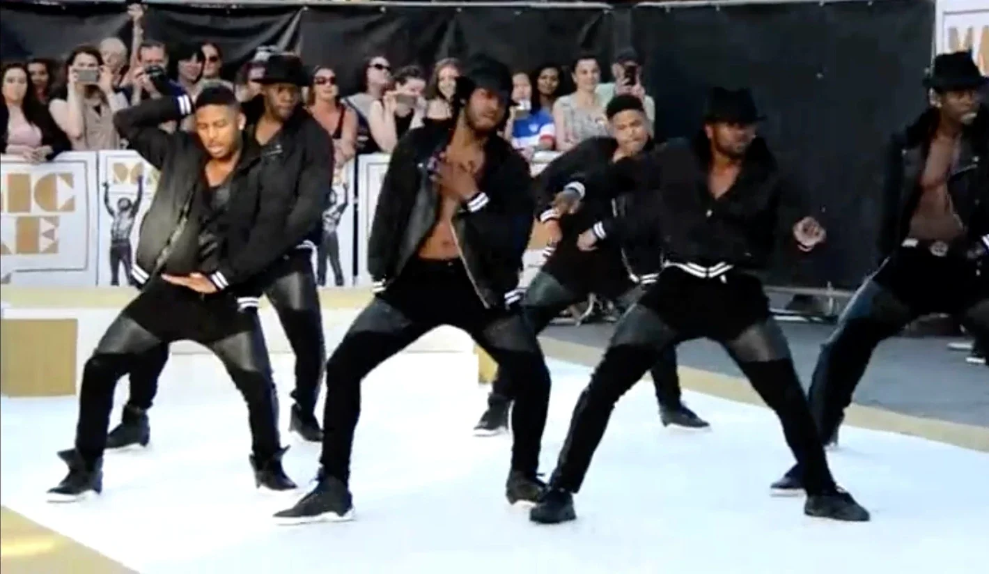 A group of male dancers performing a choreographed dance routine outdoors on a stage with an audience watching. The dancers are wearing black jackets, black pants, and black shoes.