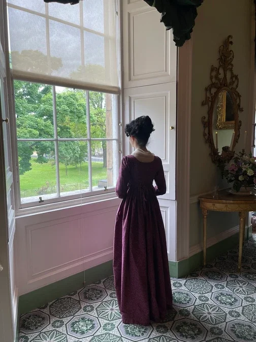 A woman in a long purple dress with dark hair styled in a regency era updo, standing inside a bright Georgian parlour, looking out a large window with multiple panes at a green park.