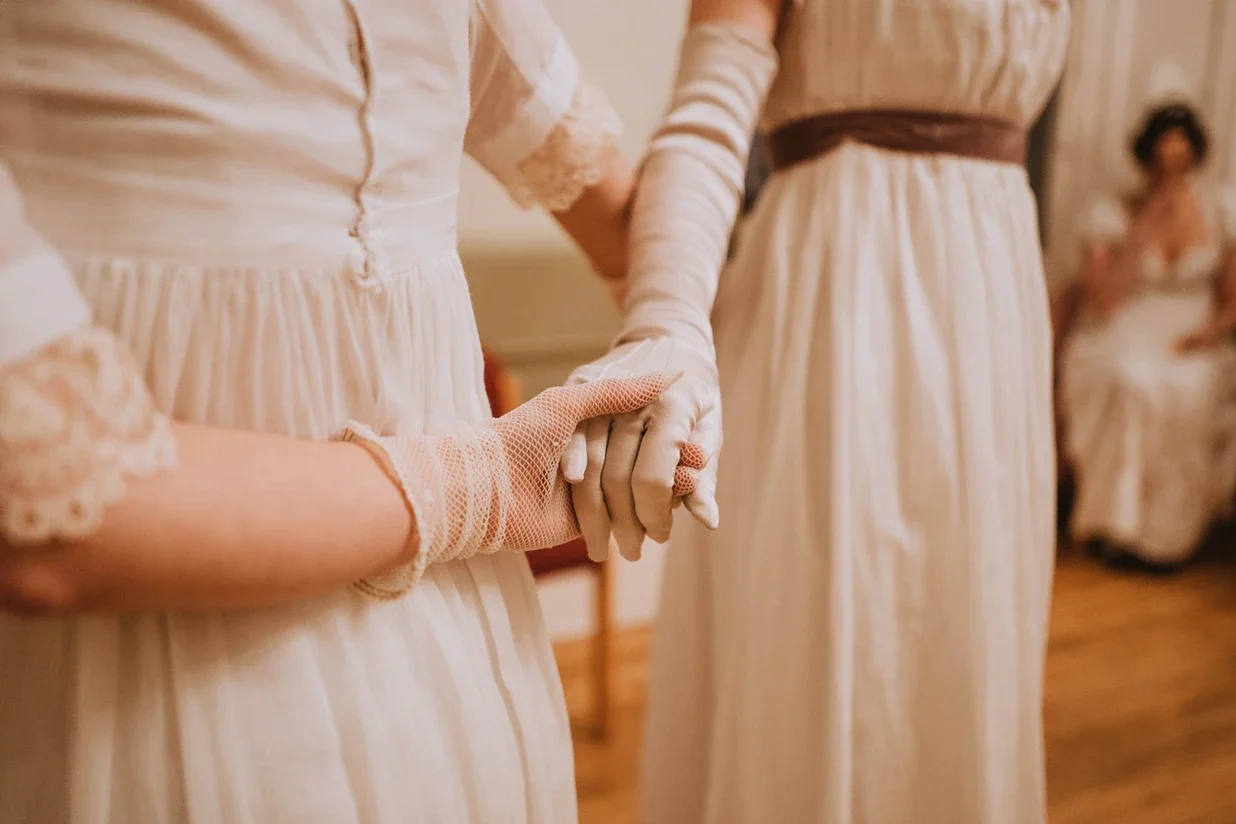 Two women wearing Regency white dresses holding hands during a dance, with other women seated in the background.