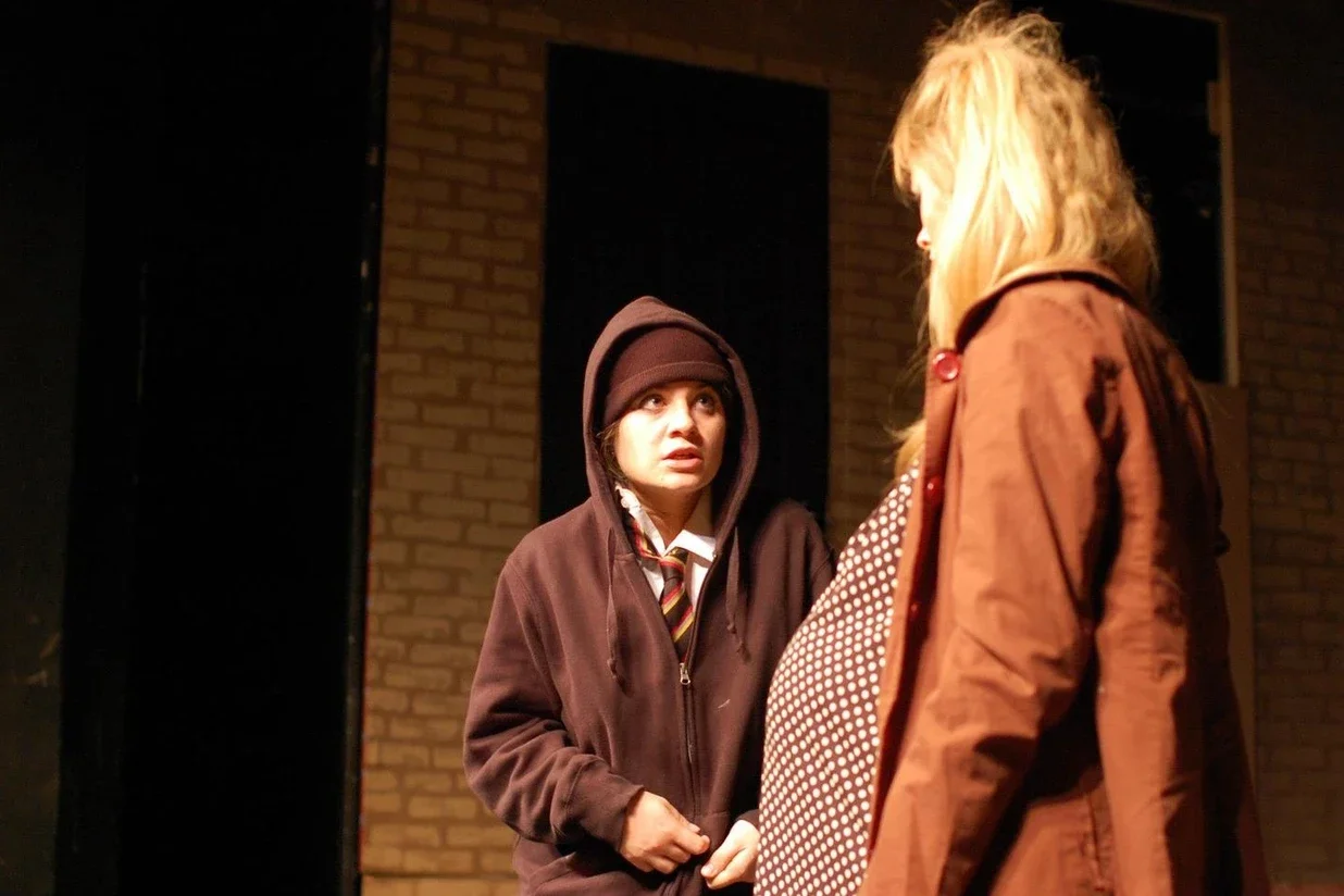Two women conversing on a stage with a brick wall background, one wearing a brown hoodie and beanie, the other dressed in a brown jacket with a polka dot shirt underneath.