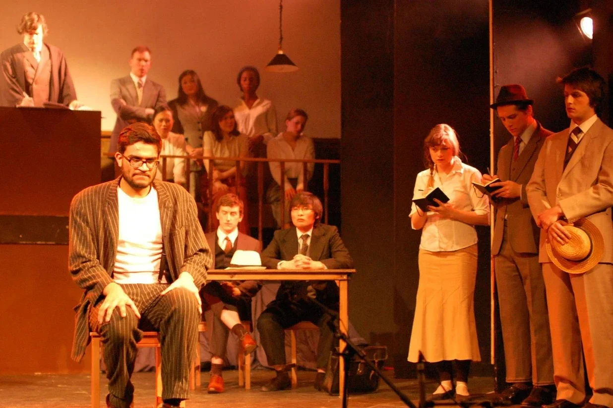The scene depicts a theatrical play with actors in period costumes. One man sits in the foreground with glasses and a striped suit, while a woman and two men stand nearby reading from notebooks. Several actors are in the background, some seated and s