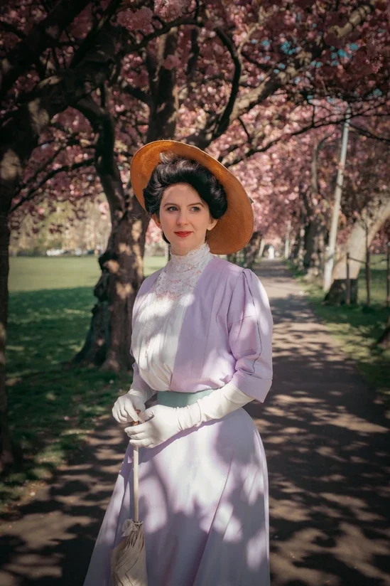 A woman dressed in Edwardian clothing, wearing a large brimmed hat, standing on a park path lined with blossoming pink cherry blossom trees.