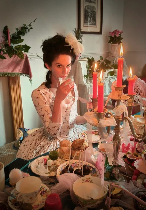 A woman dressed in Georgian floral attire, wearing a feathered headpiece, drinking from a glass at a decorated tea party with pink candles and floral arrangements.